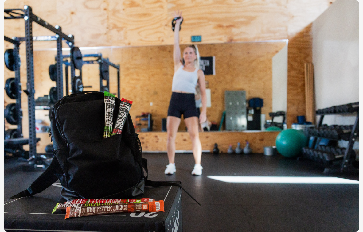 Woman wearing a tank top and shorts working out with a kettlebell with a mirror behind her and Primal Eats meat sticks tucked in the pouch of a black backpack in the foreground.